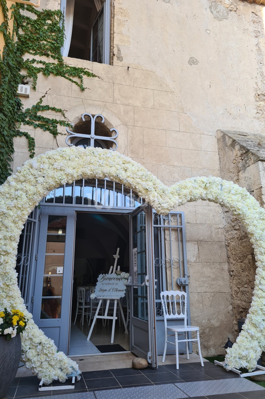 Entrée décorée d'une arche en forme de cœur composée de fleurs blanches, devant un bâtiment en pierre.