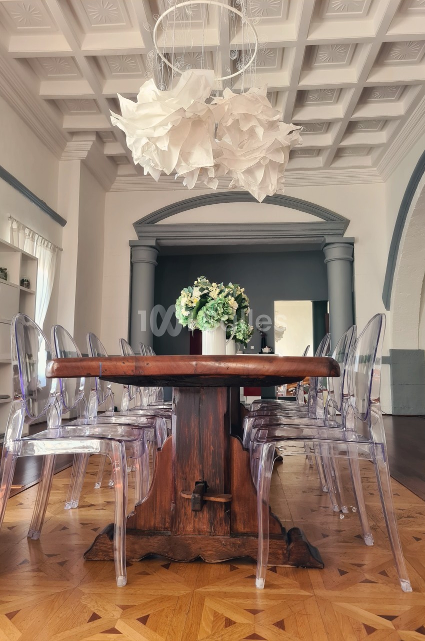 Table en bois entourée de chaises transparentes dans une pièce lumineuse avec un plafond mouluré et un lustre en papier.