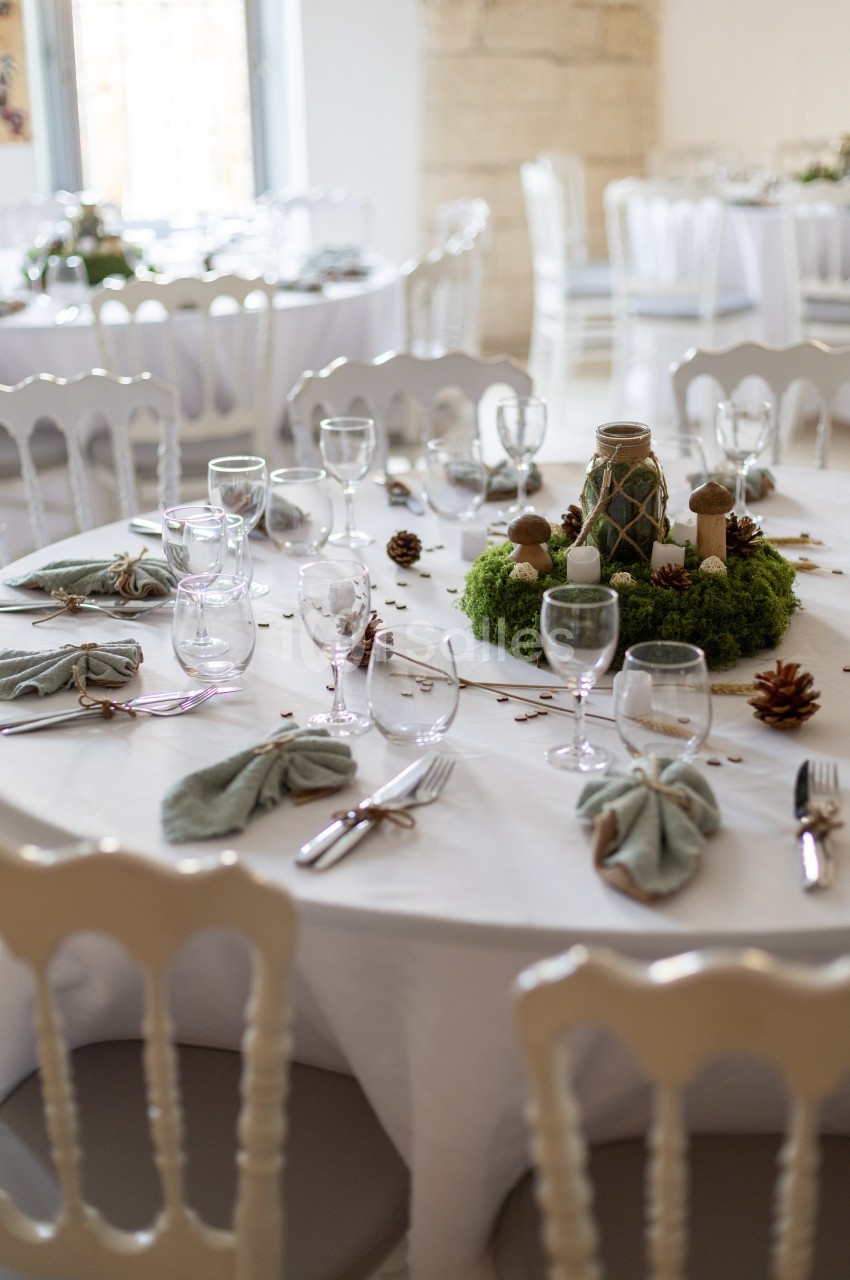Table ronde décorée pour un événement, avec nappes blanches, serviettes pliées, verres, couverts et centre de table végétal.