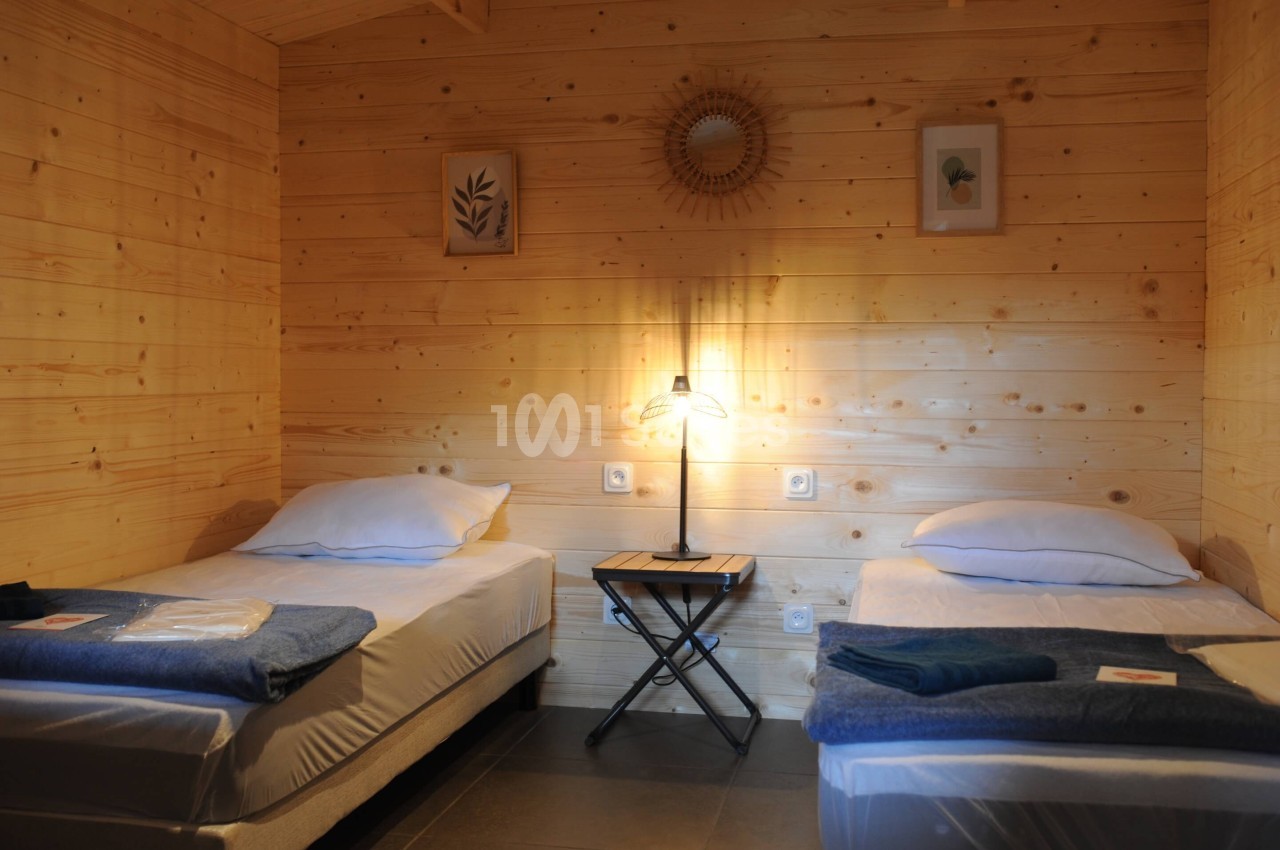 Deux lits simples avec draps bleus dans une chambre en bois clair, éclairée par une lampe sur une table centrale.
