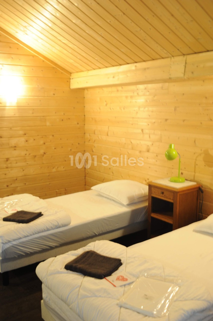 Deux lits simples avec draps blancs dans une chambre en bois clair, éclairée par une lampe murale et une lampe de chevet…