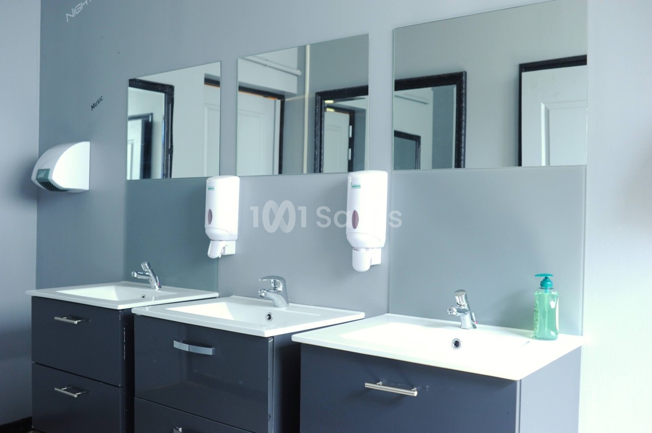 Trois lavabos modernes avec miroirs, distributeurs de savon muraux et un mur gris clair en arrière-plan.