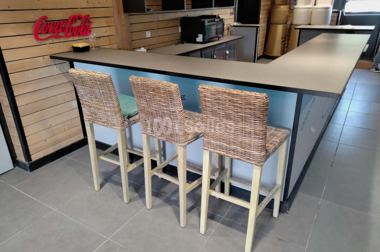 Trois tabourets en osier alignés devant un comptoir dans un espace intérieur avec murs en bois et logo Coca-Cola visible.