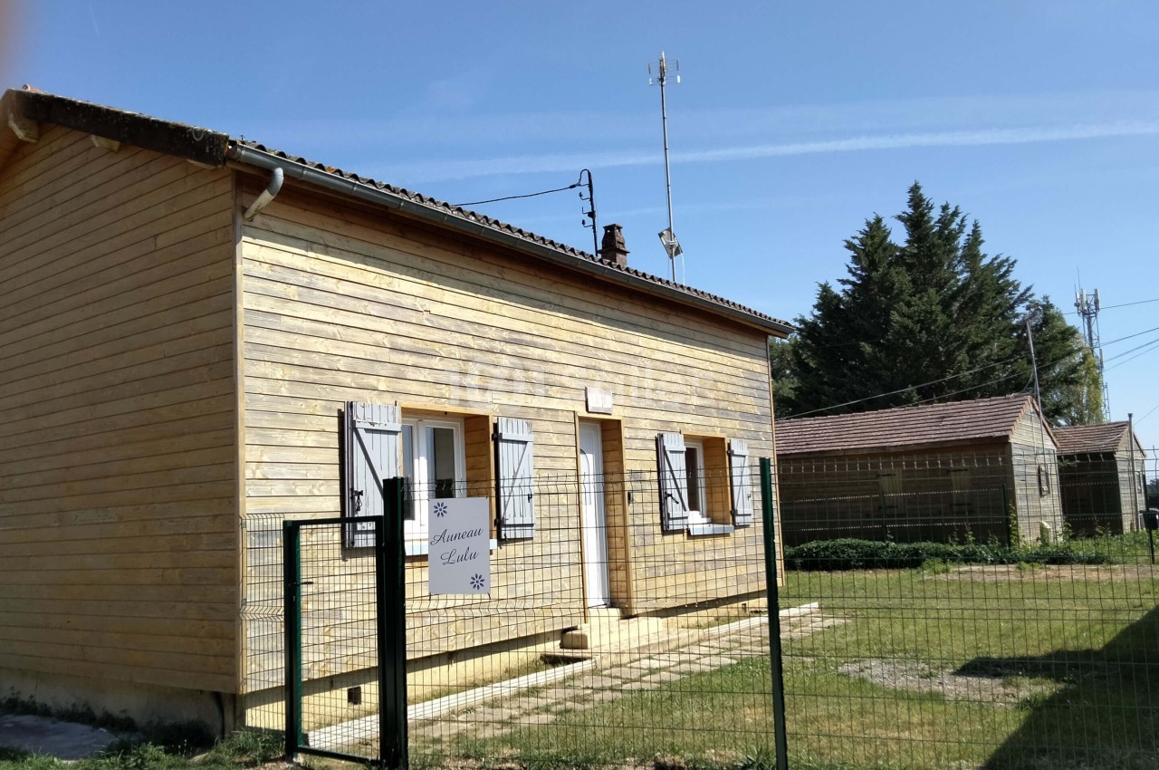 Maison en bois avec volets blancs, entourée d'une clôture, située dans un environnement rural sous un ciel dégagé.