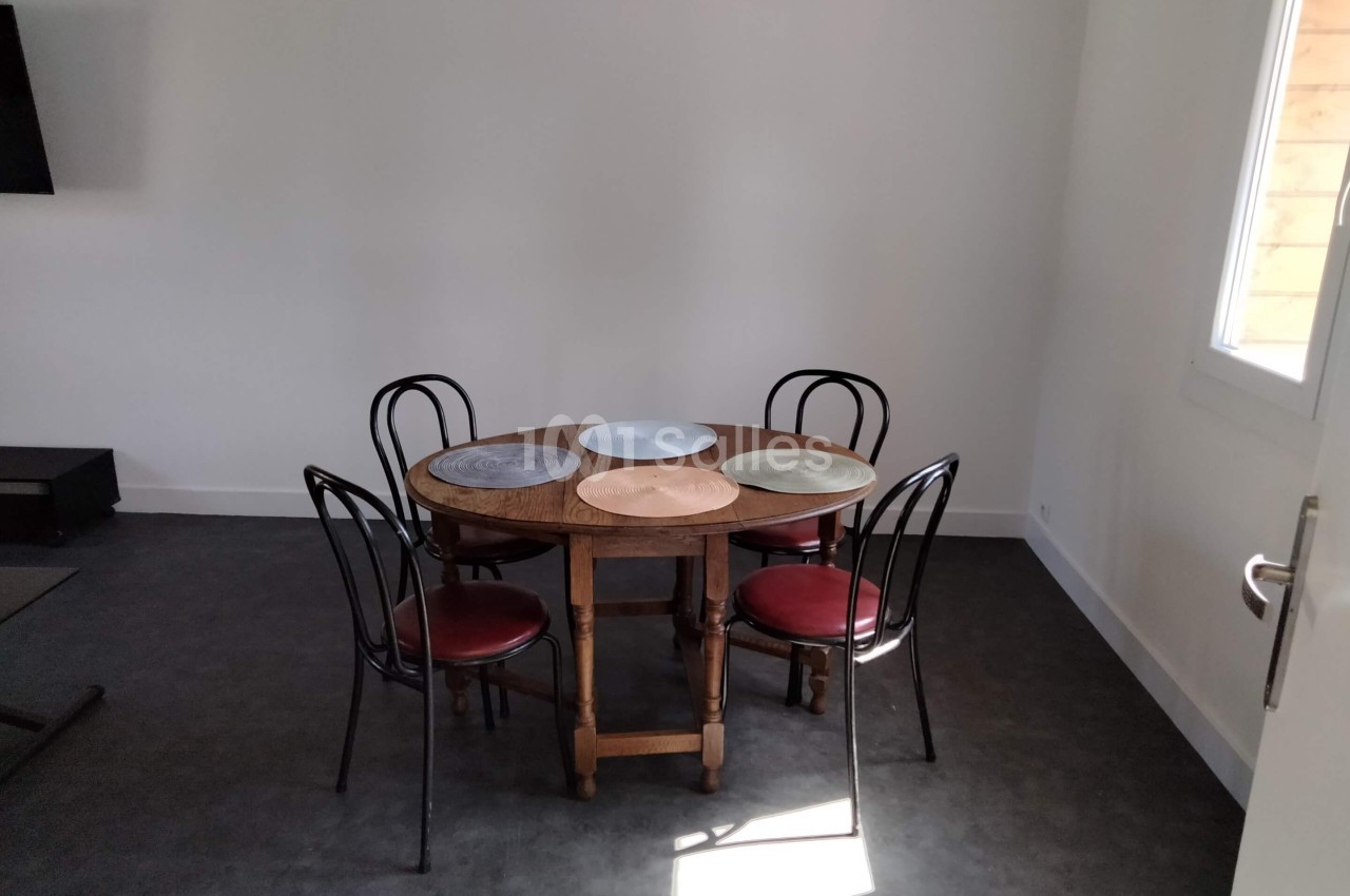 Table ronde en bois avec quatre chaises noires et des sets de table colorés dans une pièce lumineuse au sol sombre.