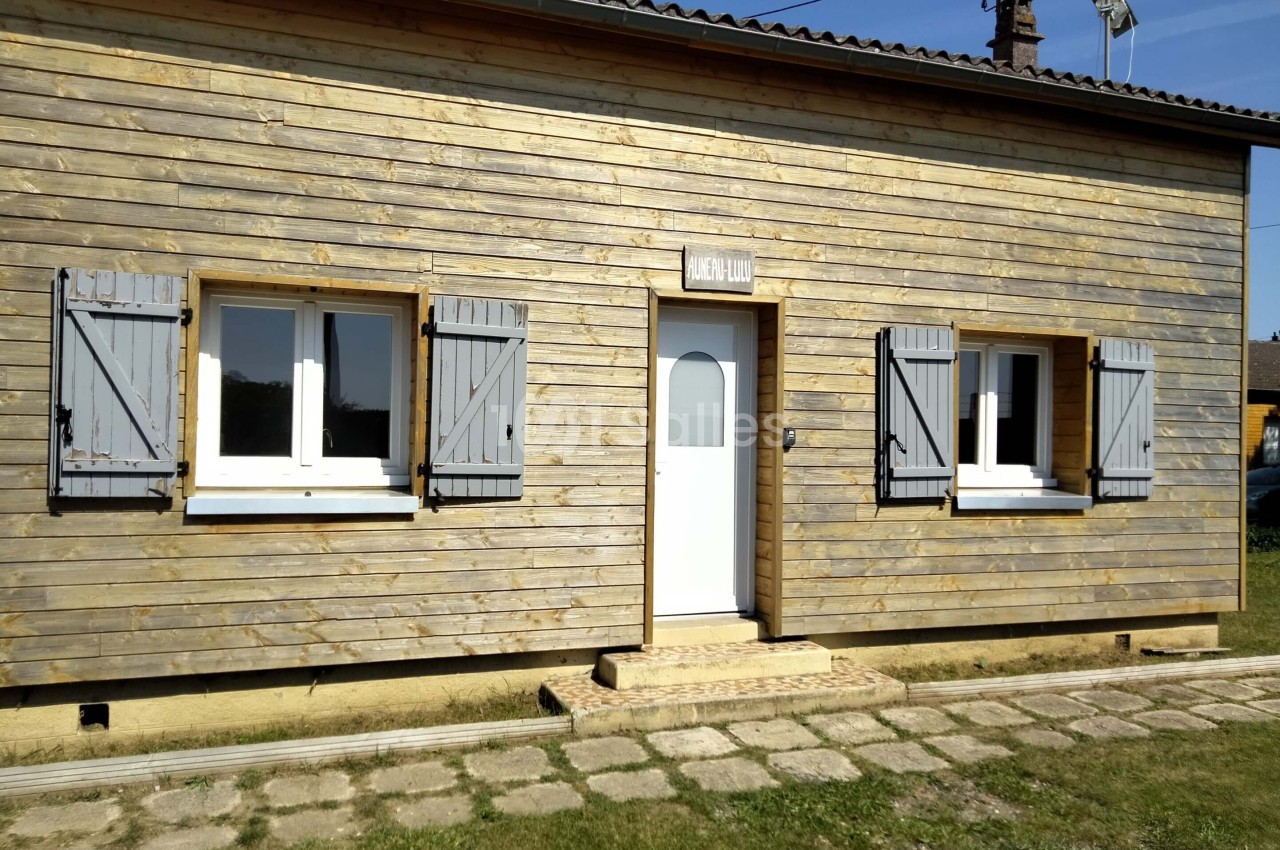 Façade d'une maison en bois avec trois fenêtres à volets gris et une porte blanche, entourée d'une pelouse.