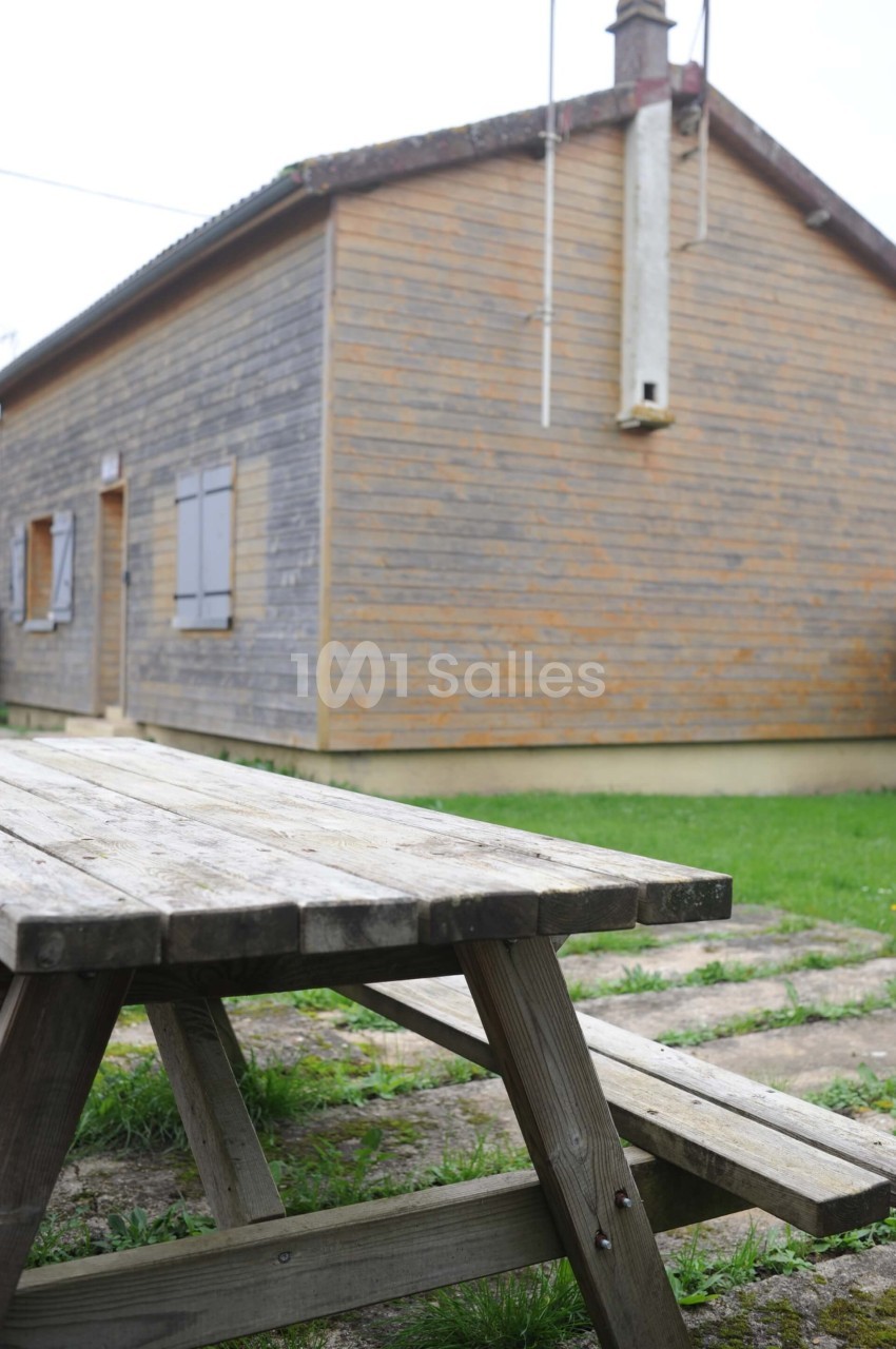 Table de pique-nique en bois devant une maison en bois avec des volets fermés, entourée de pelouse.