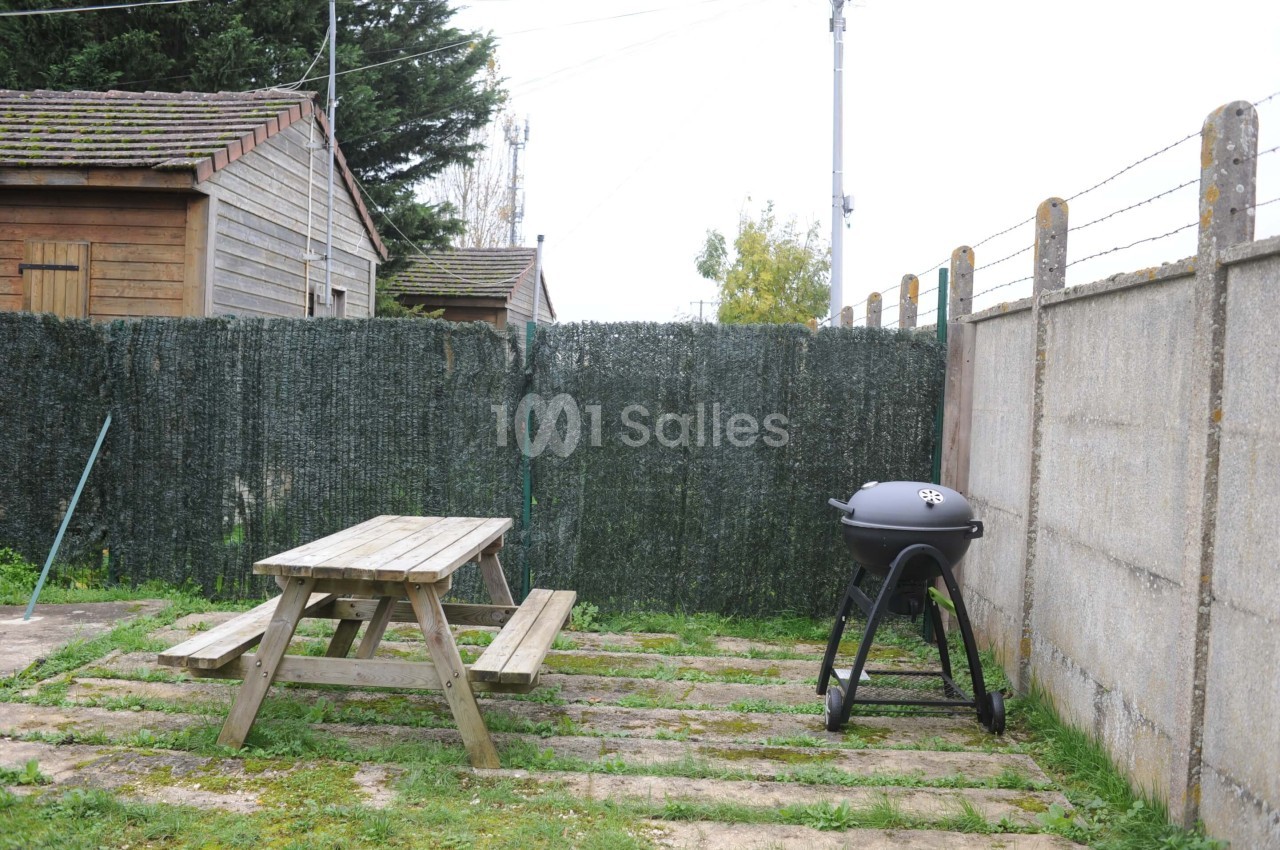 Table de pique-nique en bois et barbecue sur une terrasse extérieure entourée de clôtures et de végétation.