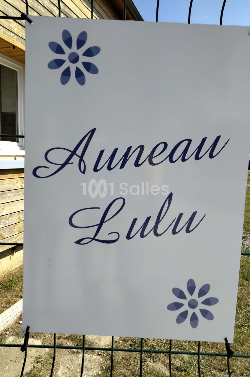 Panneau blanc avec les inscriptions ’Auneau Lulu’ en lettres bleues, décoré de motifs floraux, fixé à un grillage.