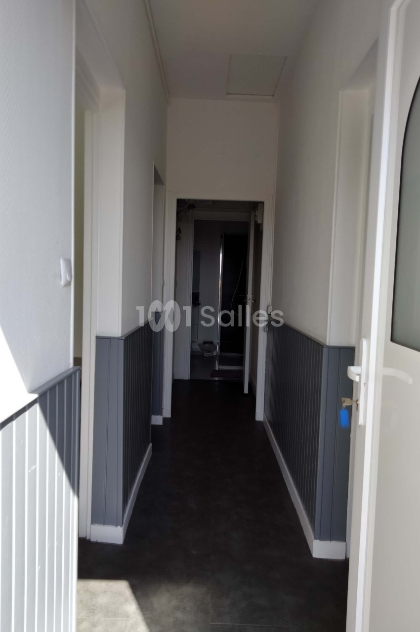 Couloir étroit avec murs blancs et gris, sol sombre, portes ouvertes et vue sur une salle de bain au fond.
