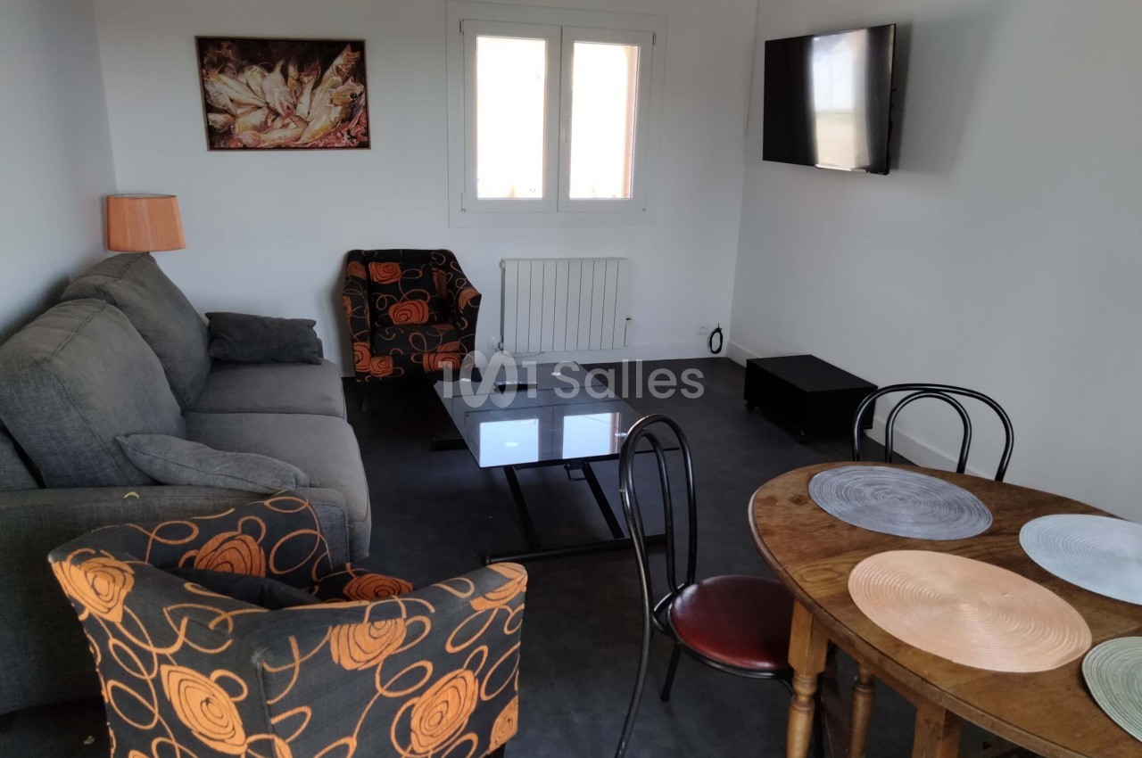 Salon avec canapé gris, fauteuils à motifs orange, table basse en verre, table ronde en bois et télévision murale.