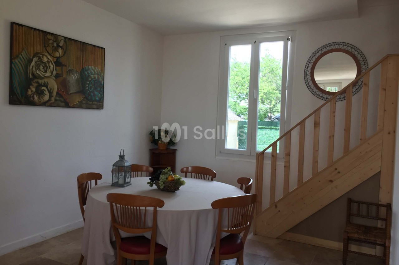 Salle à manger lumineuse avec table ronde, chaises en bois, escalier en bois et fenêtre donnant sur un jardin.