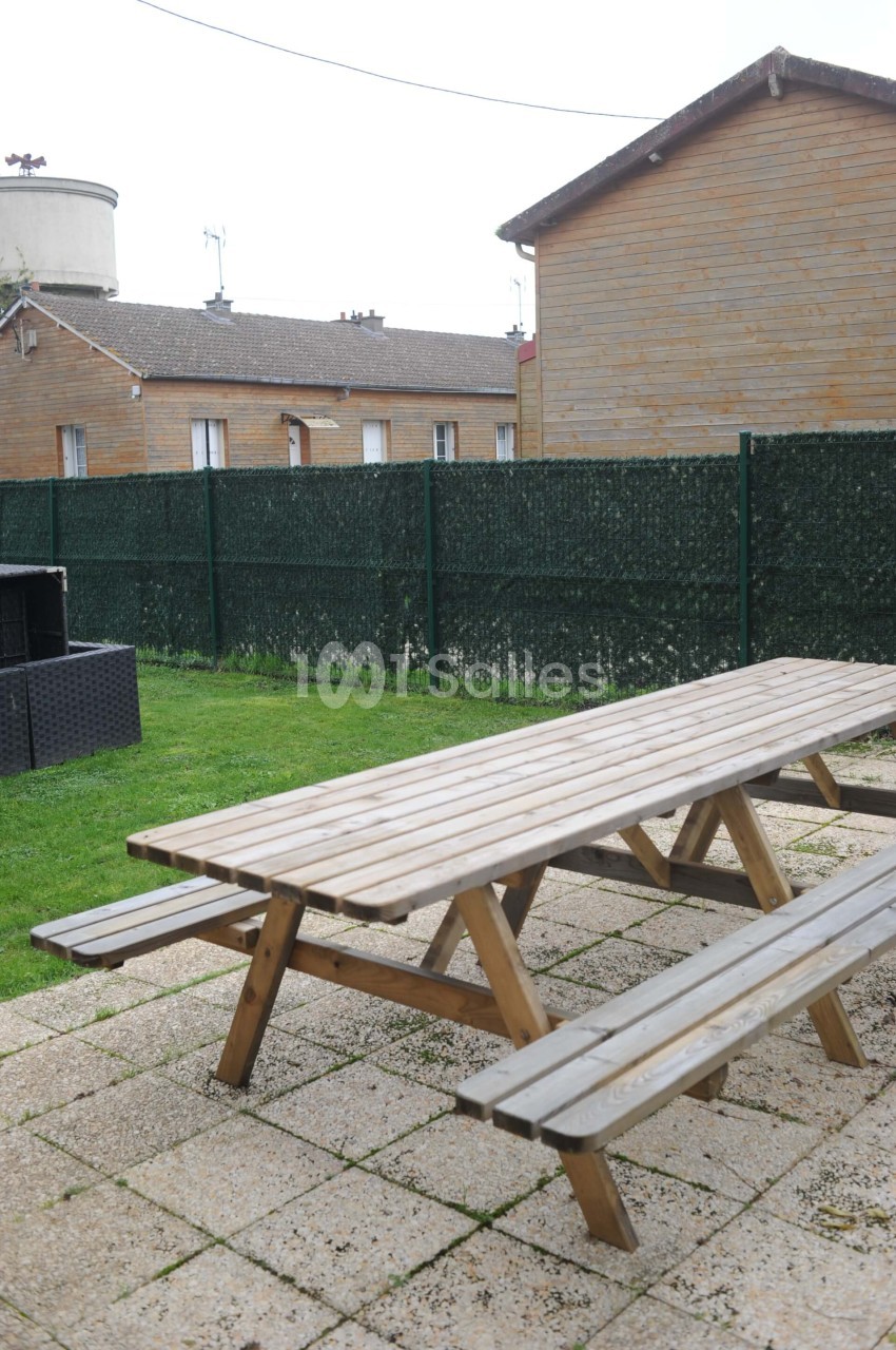 Table de pique-nique en bois sur une terrasse pavée, entourée de pelouse et clôturée par un grillage vert.