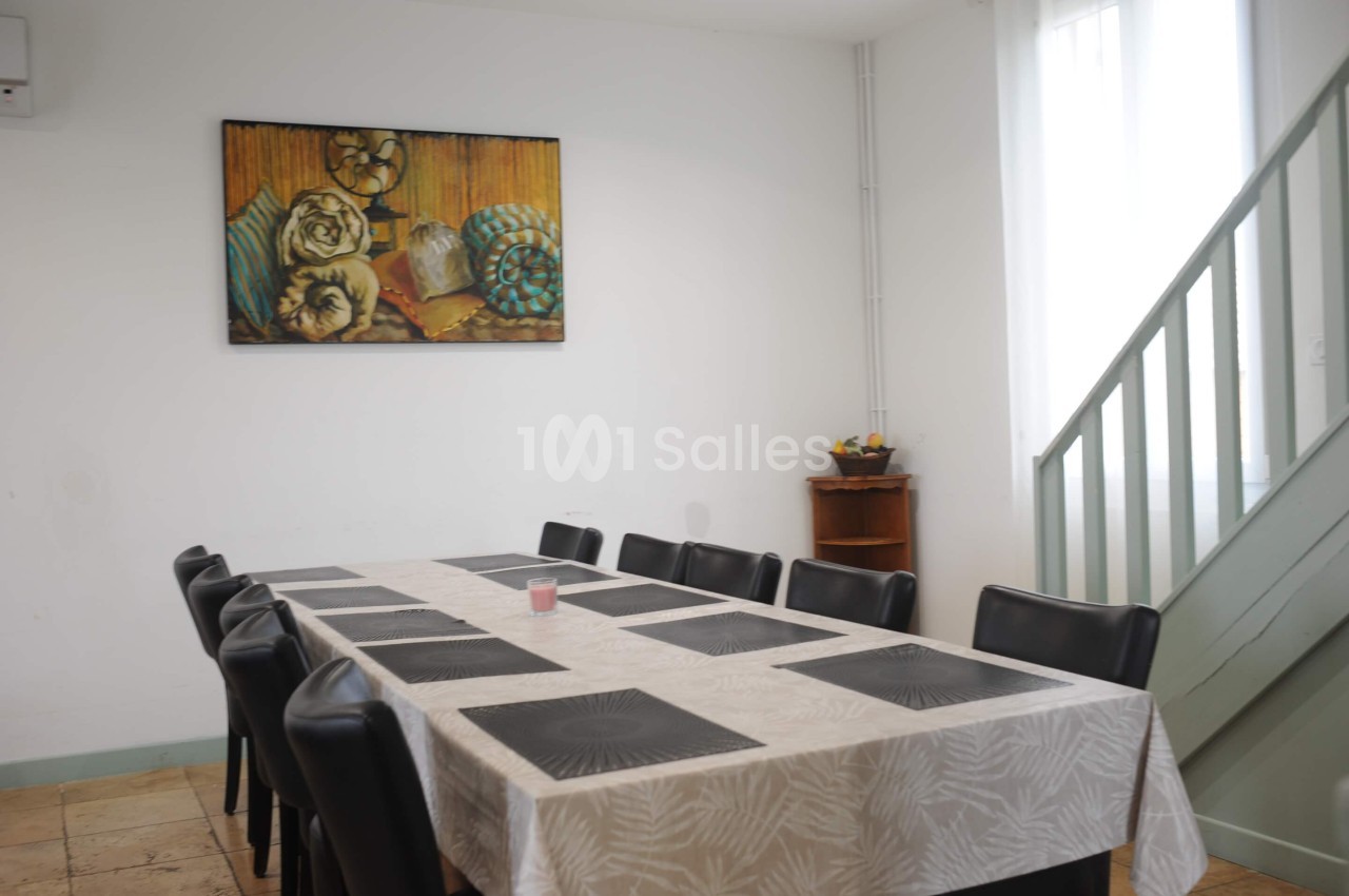Salle à manger avec une grande table rectangulaire, des chaises noires, un tableau mural et un escalier en arrière-plan.