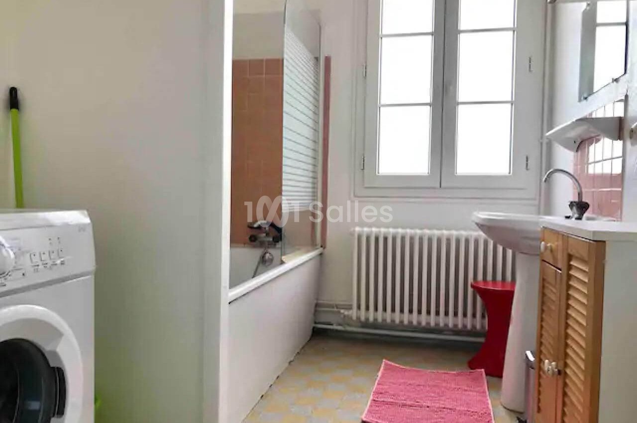 Salle de bain lumineuse avec baignoire, lavabo, machine à laver, tapis rose et fenêtre à double battant.