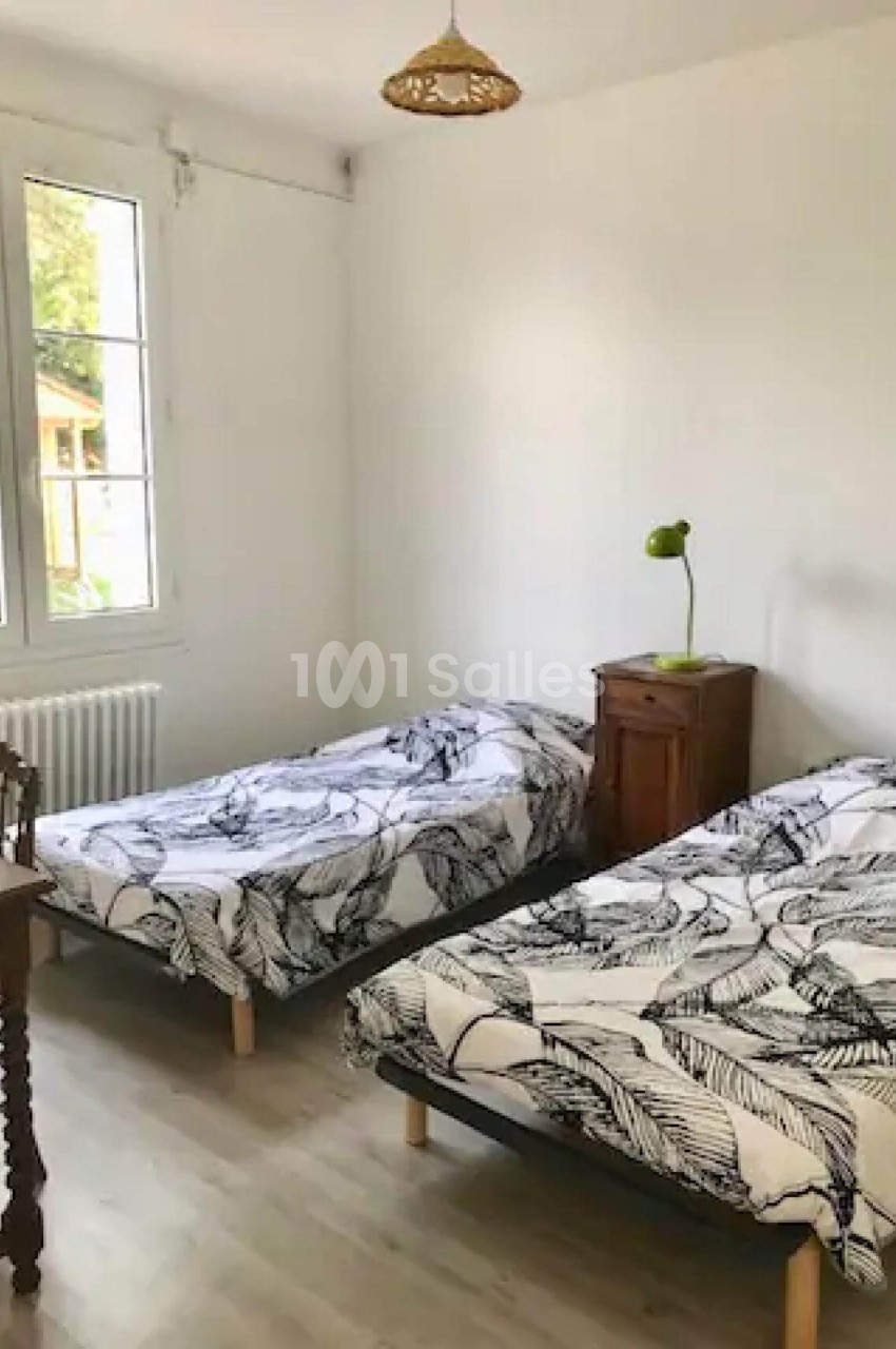 Deux lits simples avec housses à motifs noirs, une table de chevet avec lampe verte, dans une chambre lumineuse.