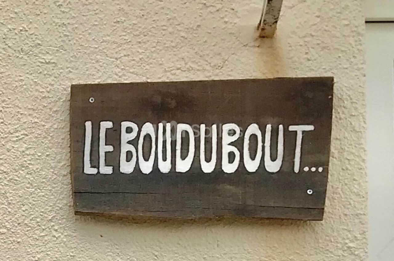 Pancarte en bois fixée sur un mur beige, portant l'inscription peinte en blanc ’LE BOUDU BOUT...’.