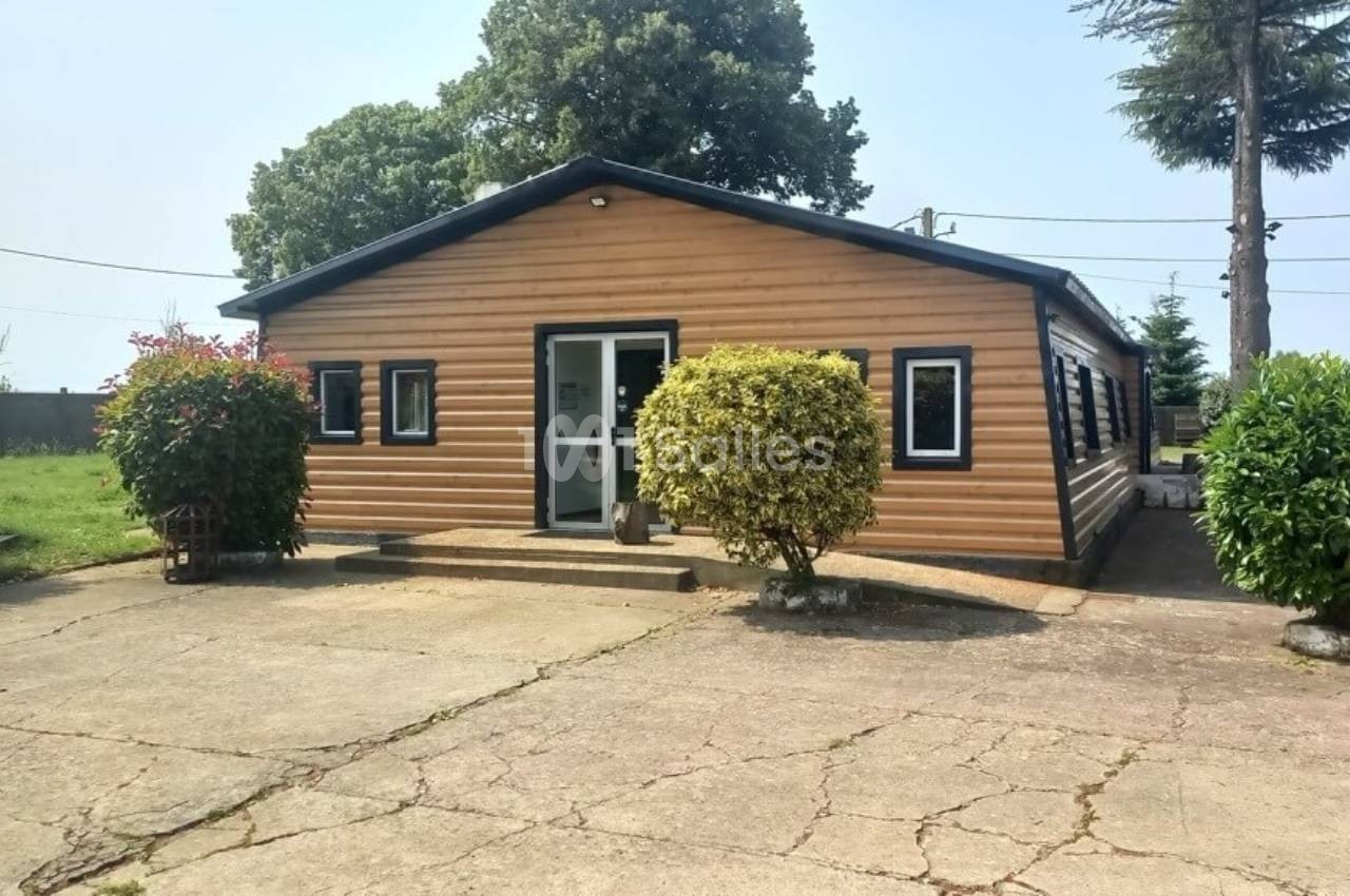 Bâtiment en bois avec plusieurs fenêtres, entouré de végétation et situé sur une surface bétonnée.