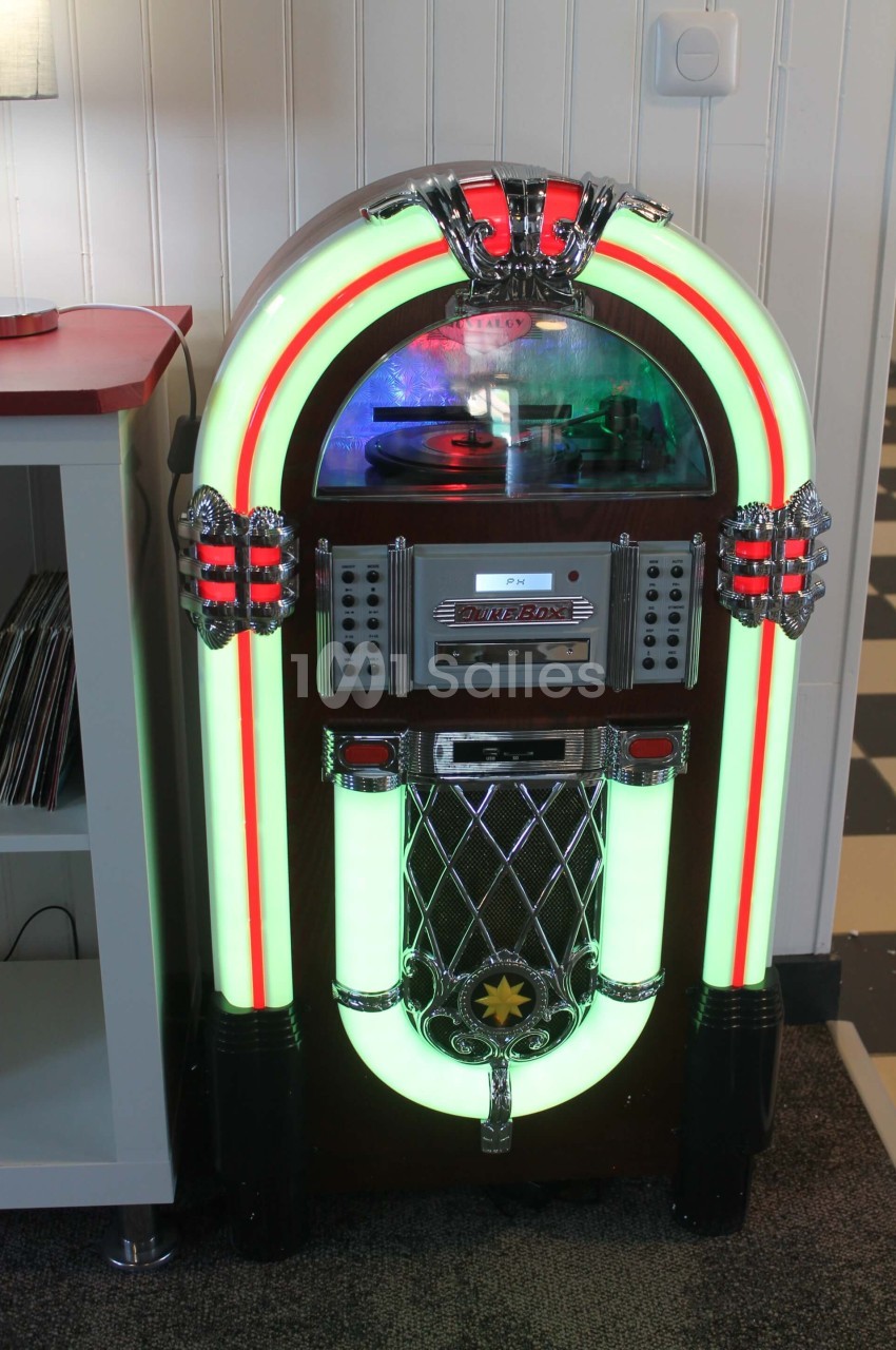 Jukebox rétro lumineux avec façade incurvée, posé sur un sol en moquette devant un mur blanc.