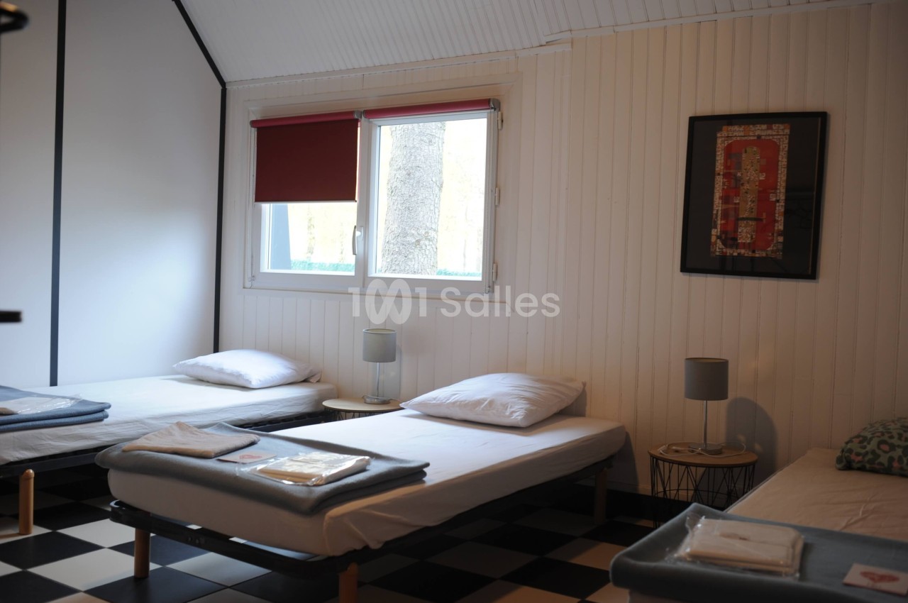 Chambre lumineuse avec trois lits simples, draps pliés, lampes de chevet et fenêtre avec store rouge.