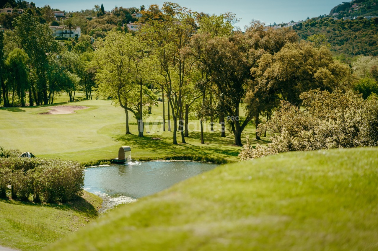Vue d'un terrain de golf verdoyant avec un étang, des arbres et des collines en arrière-plan.