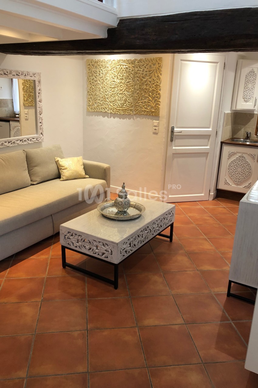 Salon lumineux avec canapé beige, table basse sculptée, miroir assorti et cuisine ouverte en arrière-plan.