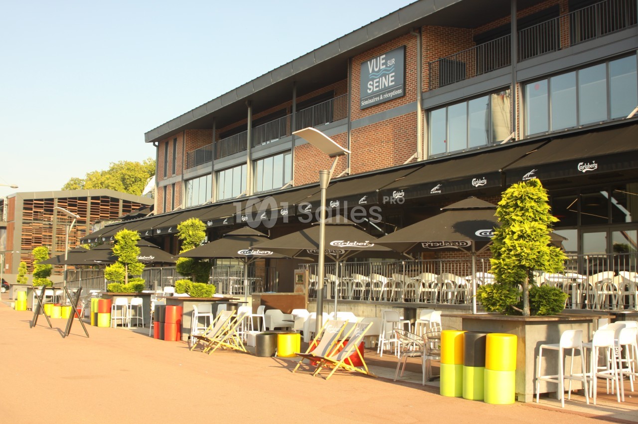 Terrasse ensoleillée avec tables, chaises et parasols devant un bâtiment en briques, bordée de plantes et de mobilier coloré.