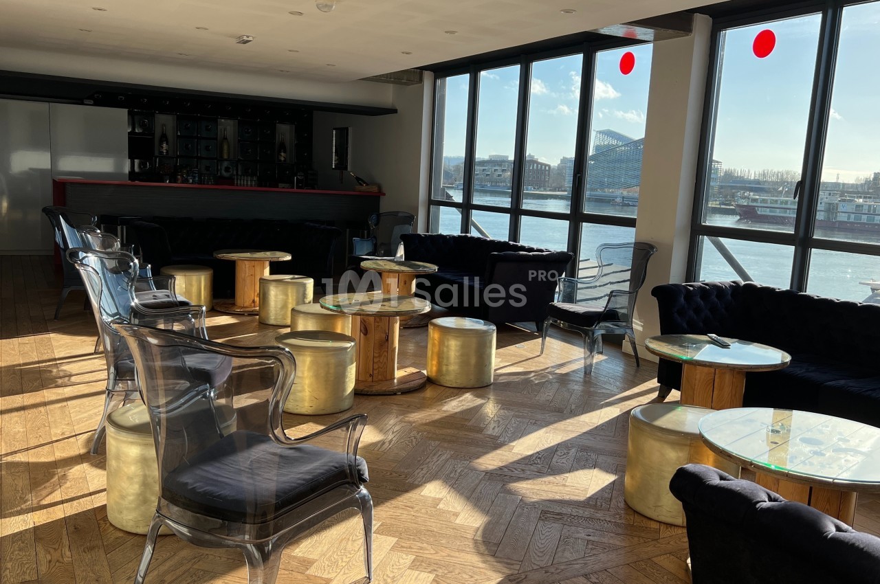 Salle lumineuse avec grandes baies vitrées, mobilier élégant en bois et métal, vue sur un fleuve et des bâtiments.