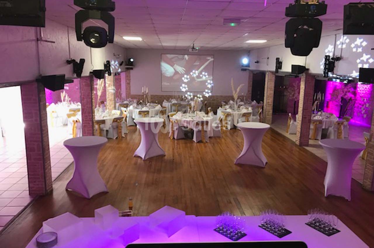 Salle de réception décorée avec des tables rondes, nappes blanches, éclairage rose et projecteurs au plafond.