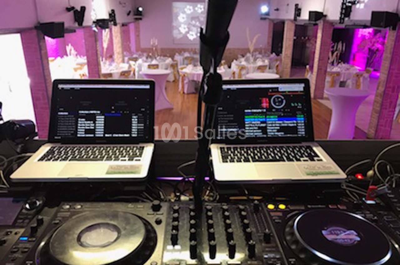 Poste de DJ avec deux ordinateurs portables, une table de mixage et une salle de réception décorée en arrière-plan.