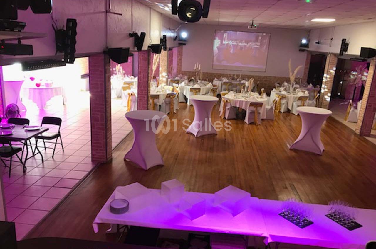 Salle de réception décorée avec tables rondes, nappes blanches, chaises ornées de rubans dorés et éclairage tamisé.