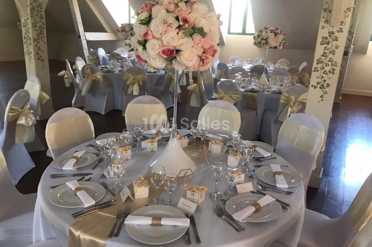 Salle de réception décorée avec des tables rondes, nappes blanches, chaises ornées de nœuds dorés et centre de table floral.