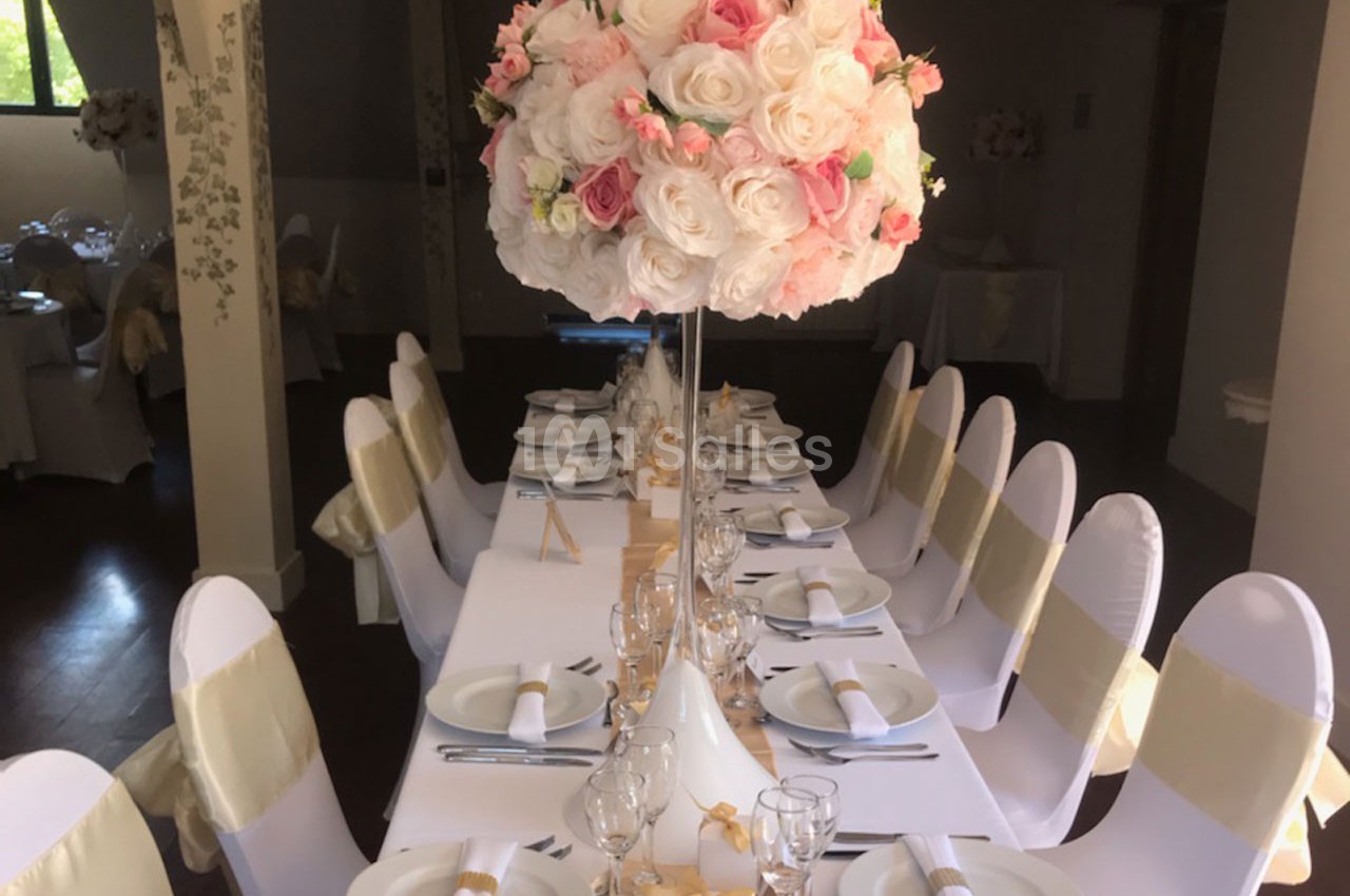 Table décorée pour un événement, avec nappes blanches, chaises ornées de rubans dorés et centre de table floral.