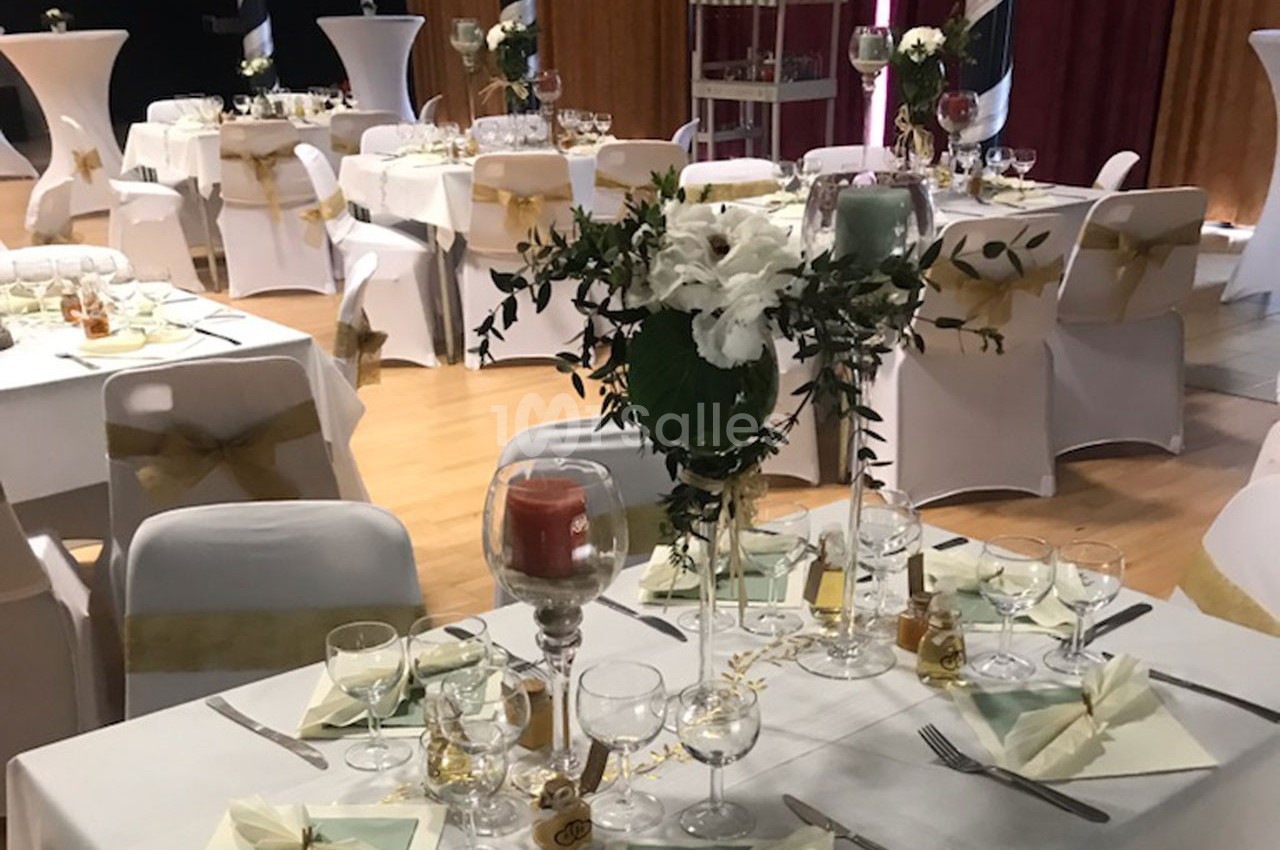 Salle décorée pour un événement, avec tables dressées, nappes blanches, centres de table fleuris et bougies.