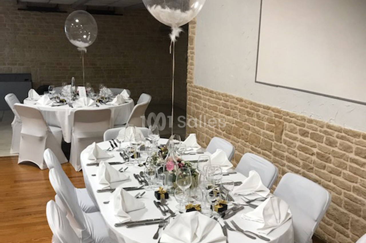 Salle décorée pour un événement avec tables rondes dressées, nappes blanches, chaises houssées et ballons transparents.