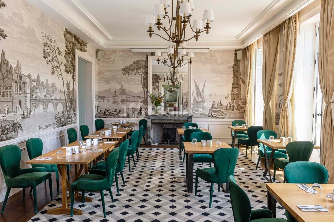 Salle de restaurant lumineuse avec des chaises vertes, tables en bois, fresques murales et un lustre central.