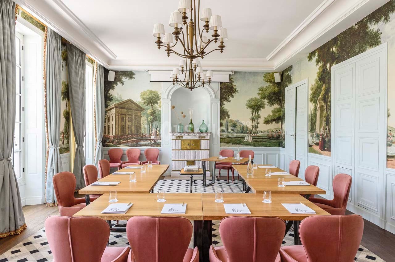 Salle de réunion lumineuse avec tables en bois, chaises roses, fresques murales et lustre central élégant.