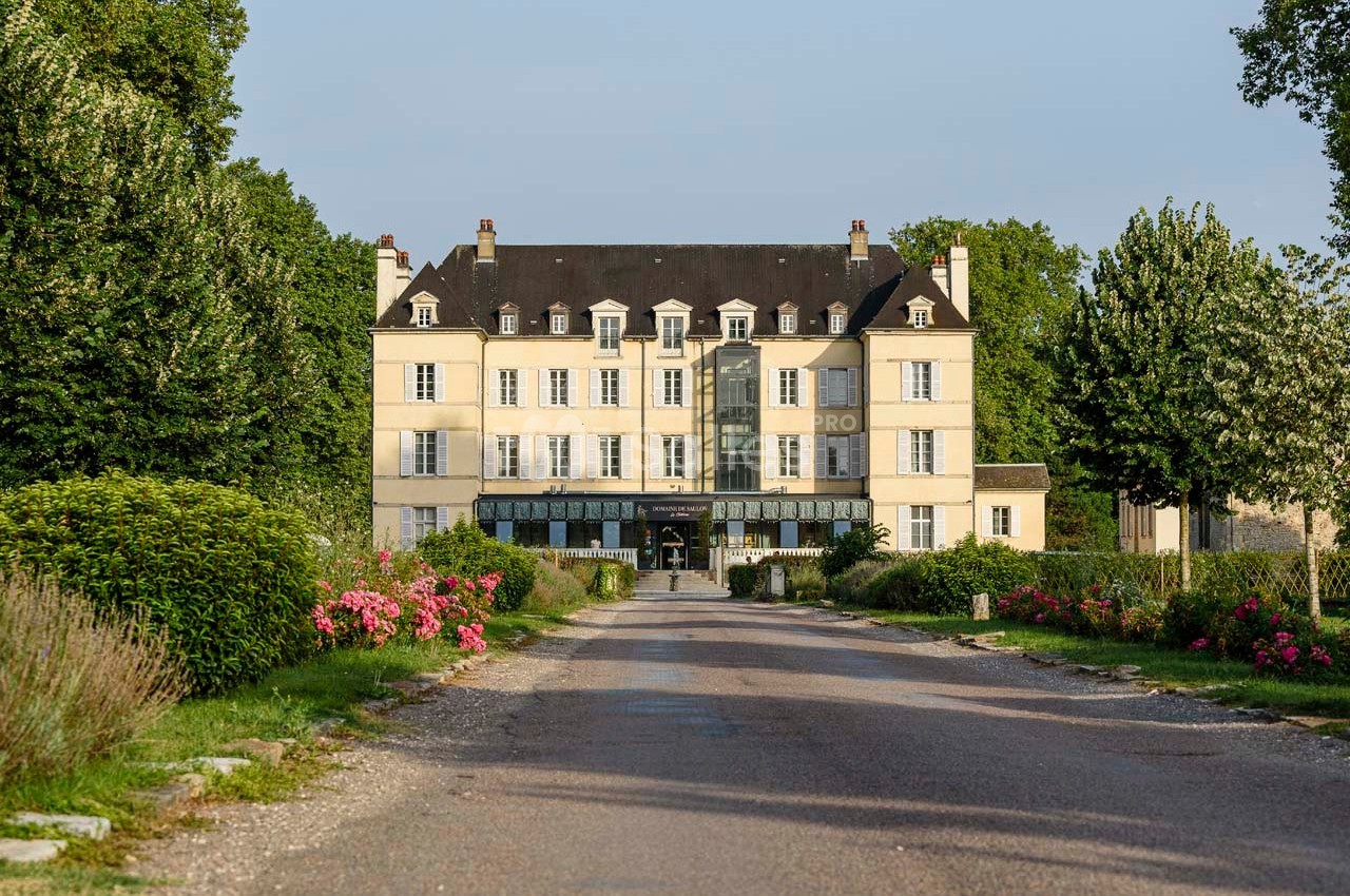 Façade d'un grand bâtiment de style classique entouré de jardins fleuris et d'une allée bordée d'arbres.
