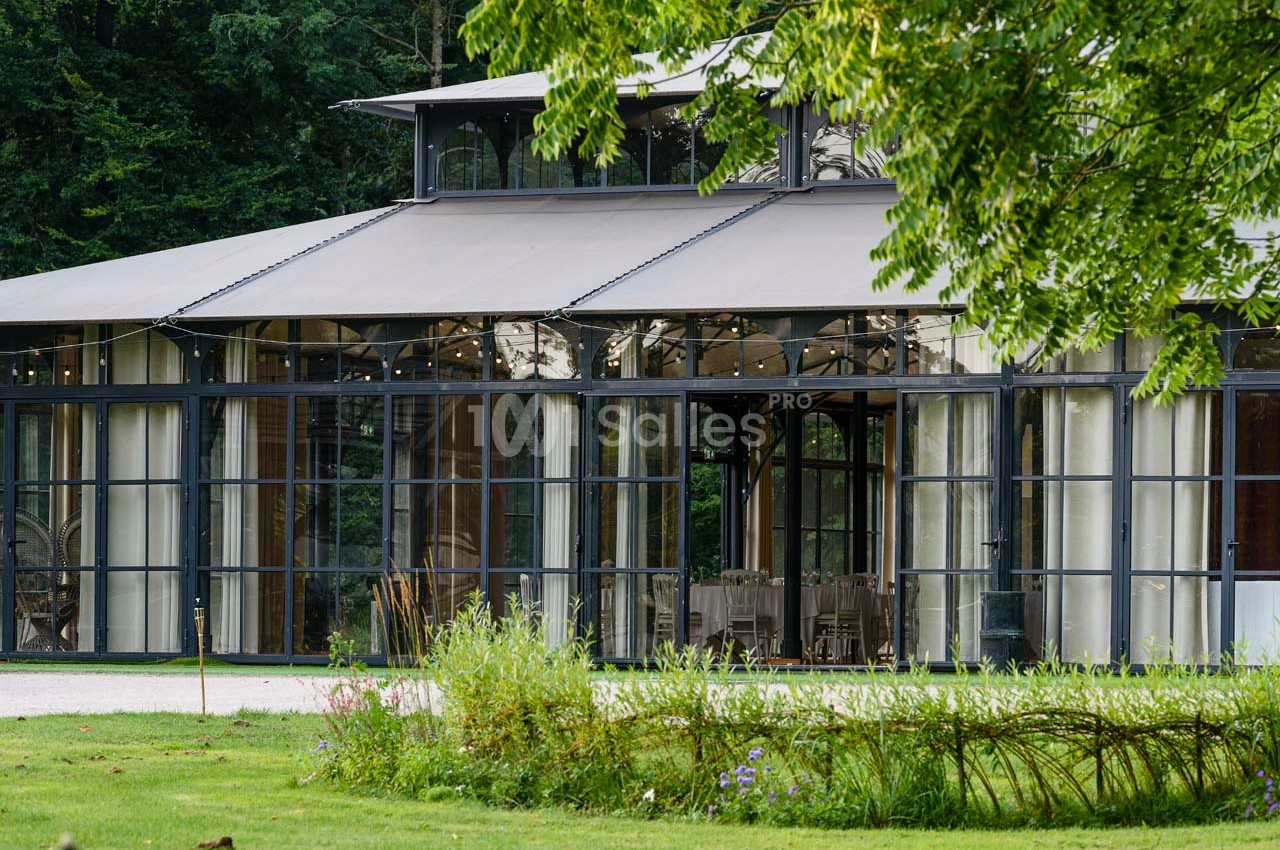 Vue extérieure d'une verrière moderne entourée de verdure, avec des tables dressées visibles à l'intérieur.