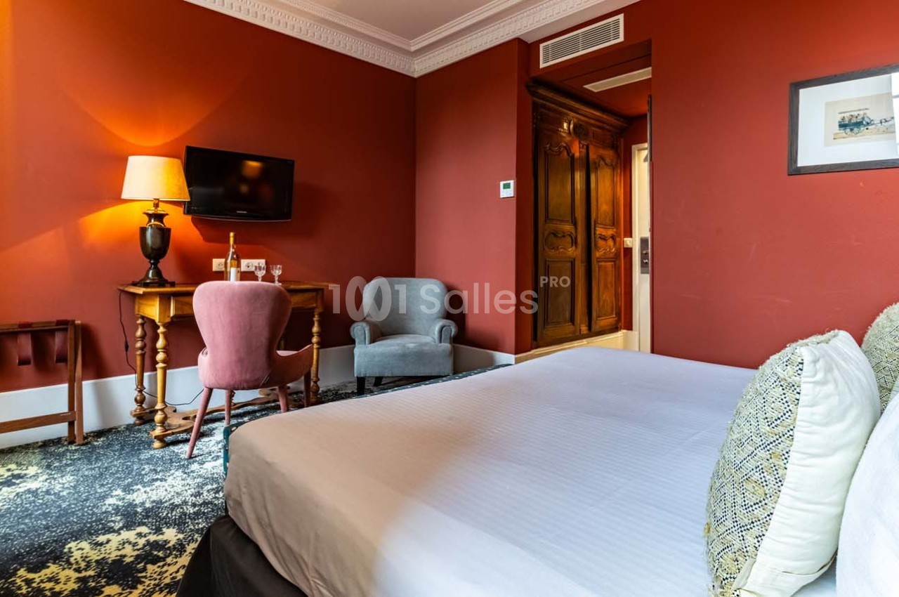 Chambre d'hôtel avec lit double, bureau en bois, fauteuils, mur rouge et éclairage chaleureux.