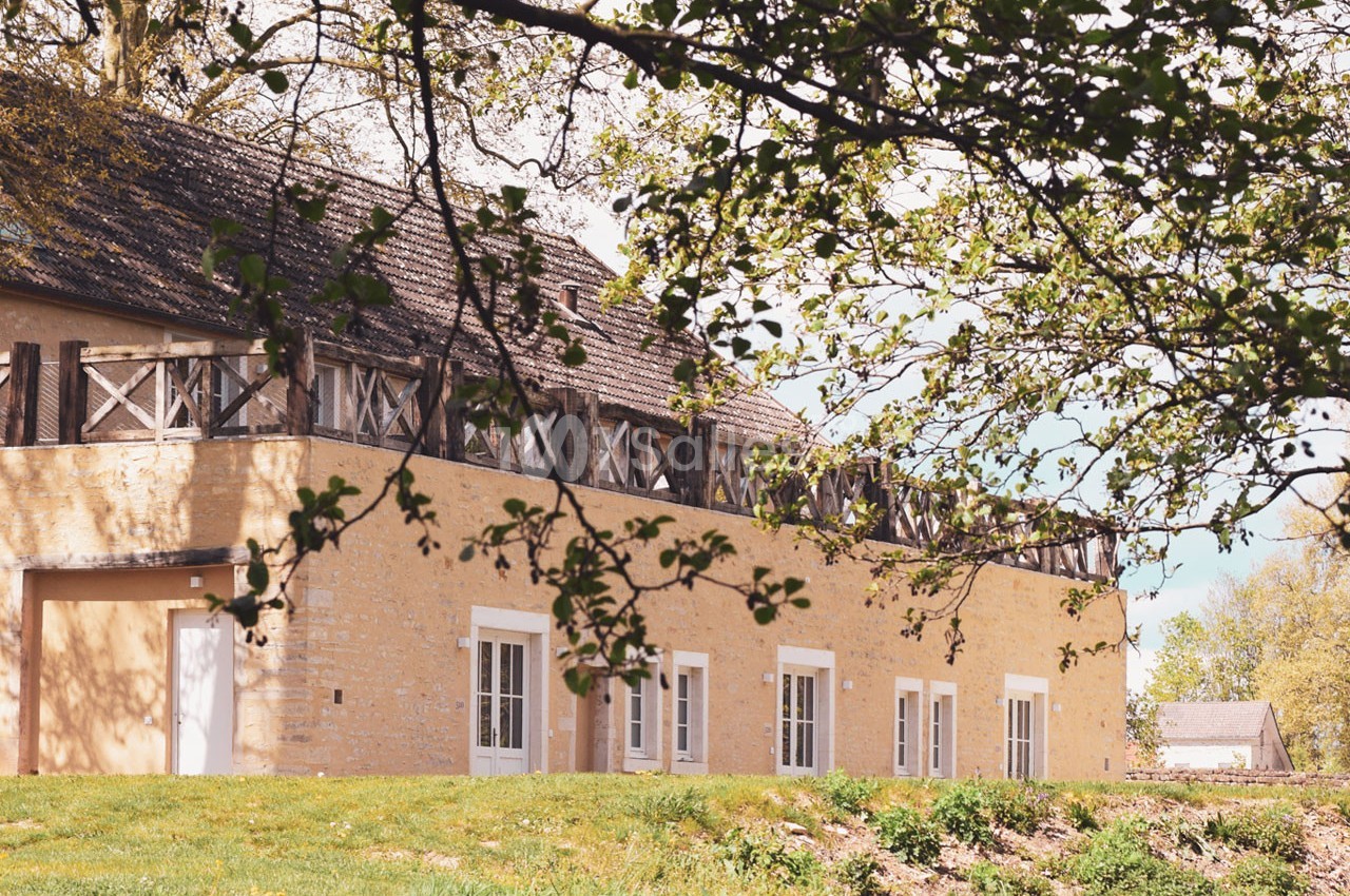 Bâtiment en pierre beige avec fenêtres blanches, entouré de verdure et partiellement ombragé par des branches d'arbre.