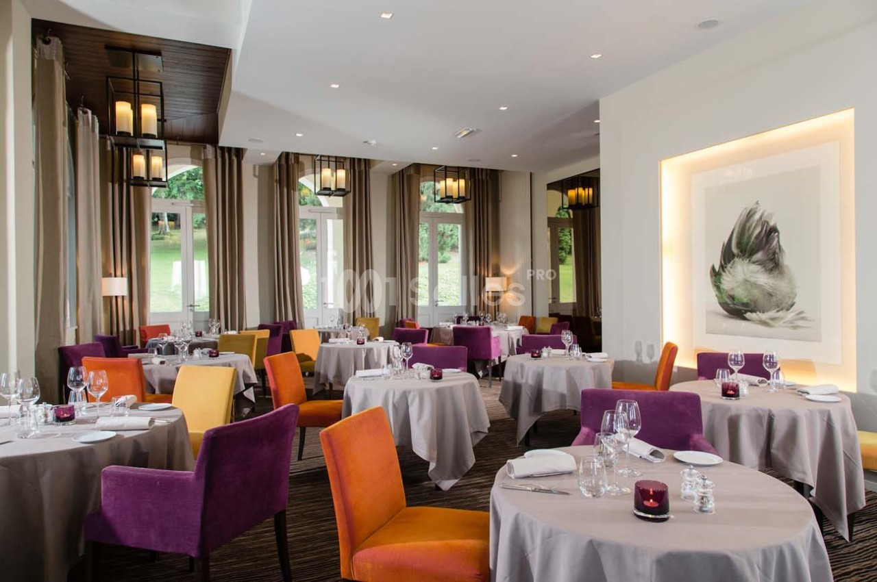 Salle de restaurant lumineuse avec tables dressées, chaises colorées et grande baie vitrée donnant sur un espace vert.