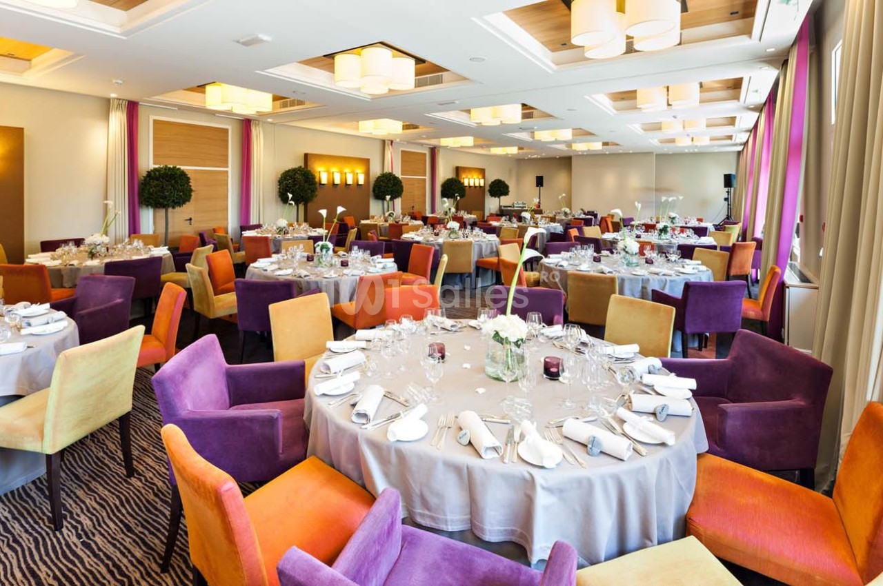 Salle de réception lumineuse avec tables rondes dressées, chaises colorées et décoration élégante.