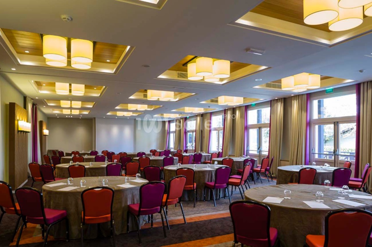 Salle de conférence lumineuse avec des tables rondes dressées, des chaises rouges et de grandes fenêtres.