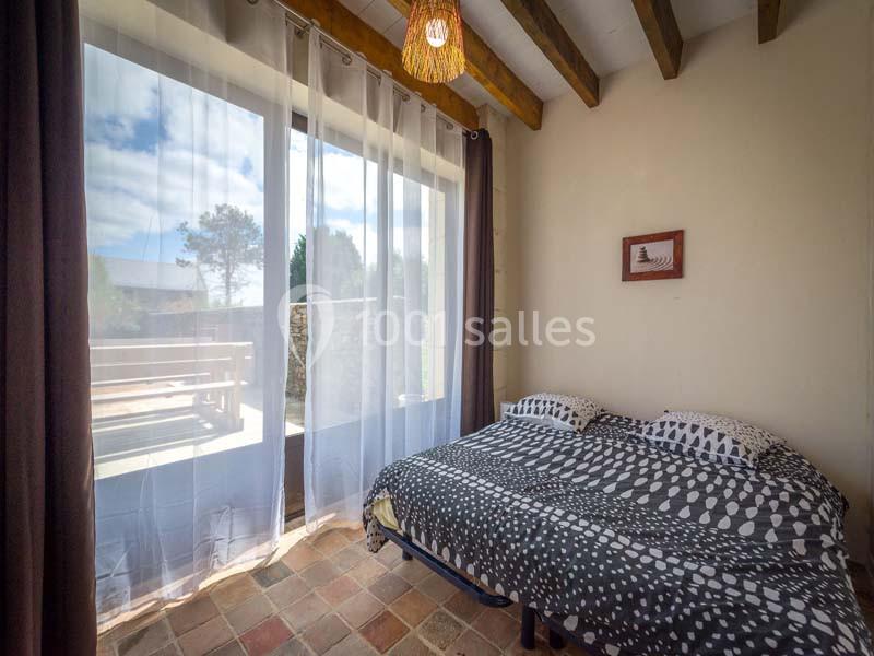 Chambre lumineuse avec lit double, rideaux transparents, plafond en poutres apparentes et vue sur une terrasse ensoleillée.