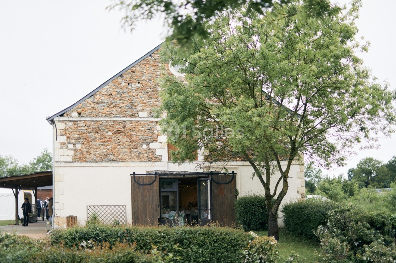 Façade d'une maison en pierre avec un arbre à droite, entourée de verdure et d'arbustes.