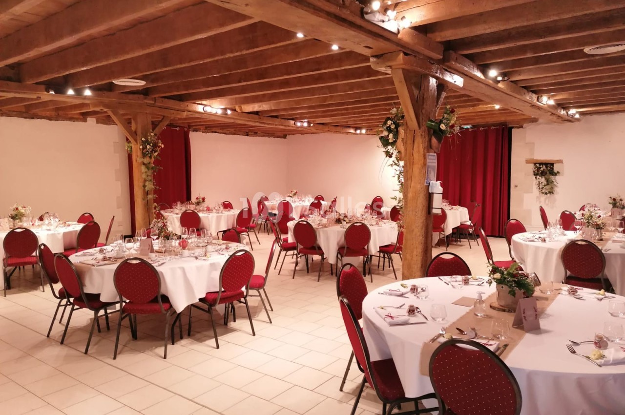 Salle de réception décorée avec des tables rondes dressées, chaises rouges et poutres apparentes en bois.