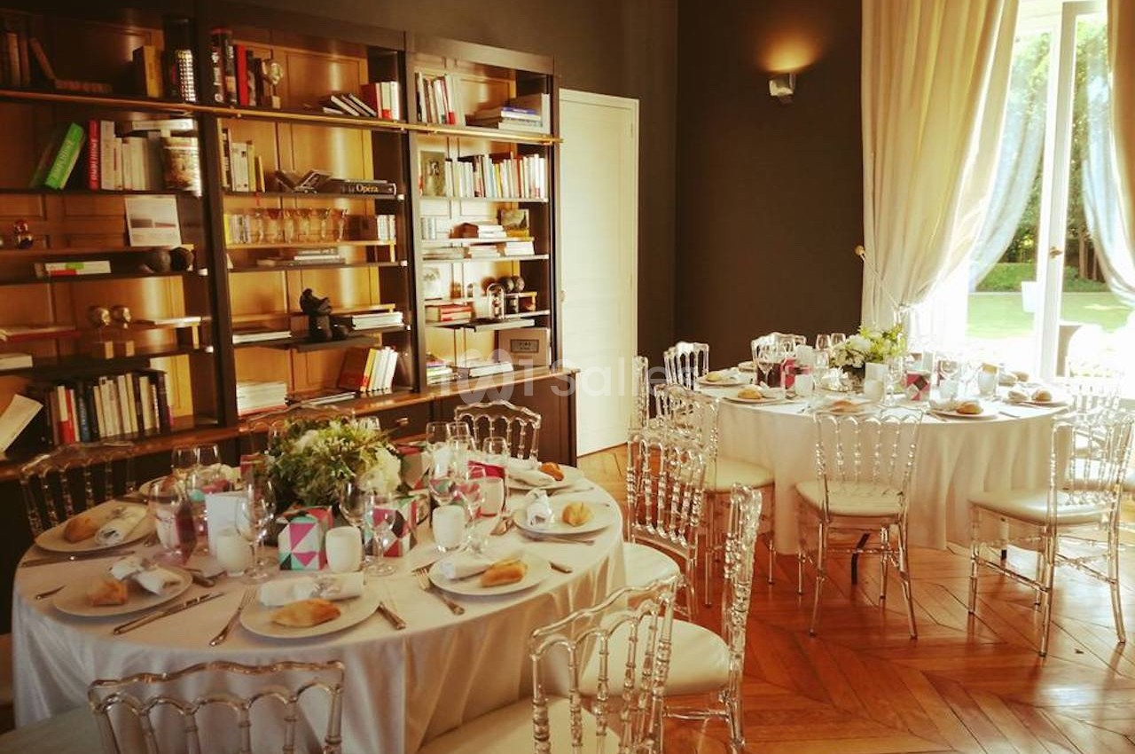 Salle élégante avec tables rondes dressées pour un repas, chaises transparentes et bibliothèque en arrière-plan.