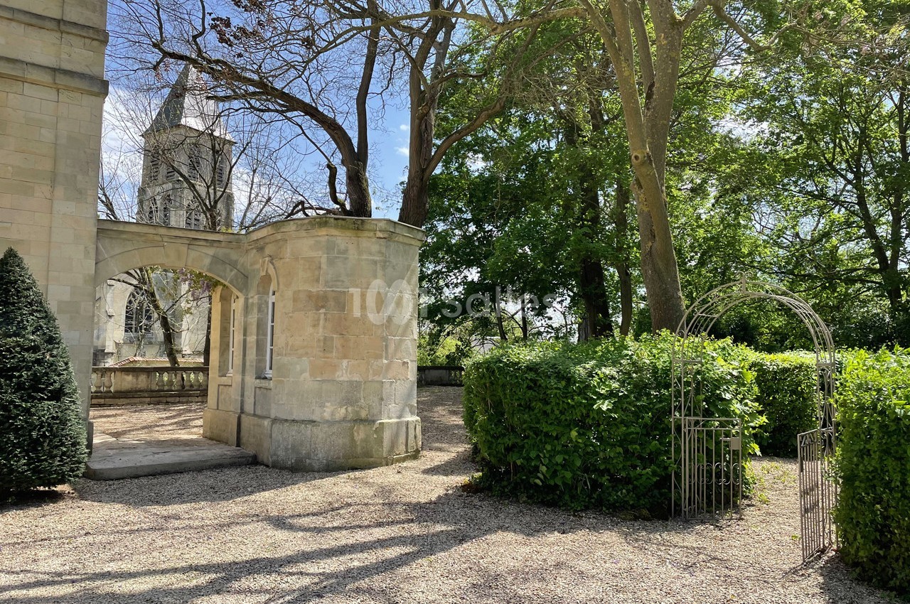 Allée gravillonnée bordée de haies et d'arbres, avec une arche métallique et un bâtiment en pierre à gauche.