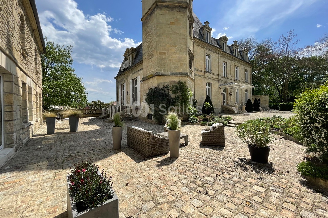 Cour pavée avec mobilier d'extérieur, entourée d'un bâtiment en pierre et de végétation sous un ciel partiellement nuageux.